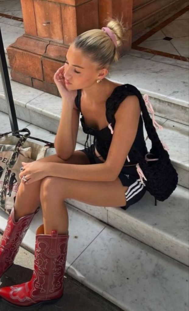 A person sitting on stairs, wearing red cowboy boots, black sports shorts, and a black top, with a hair bun and holding their chin in thought.