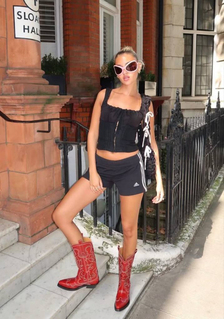 A woman poses on the steps of a building, wearing red cowboy boots, a black crop top, and black athletic shorts, paired with oversized sunglasses and a bag.