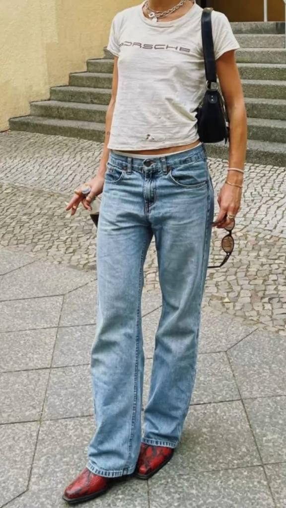 A person wearing a cropped t-shirt and light blue flared jeans, complemented by stylish cowboy boots, stands on a cobblestone surface near a staircase.