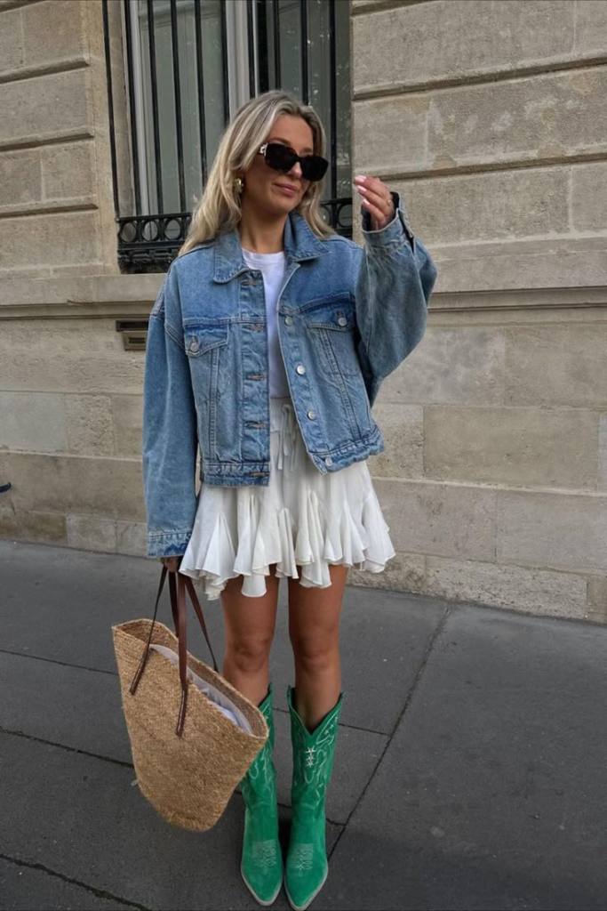 A fashionable woman wearing a denim jacket, white mini skirt, and green cowboy boots, holding a straw tote bag.