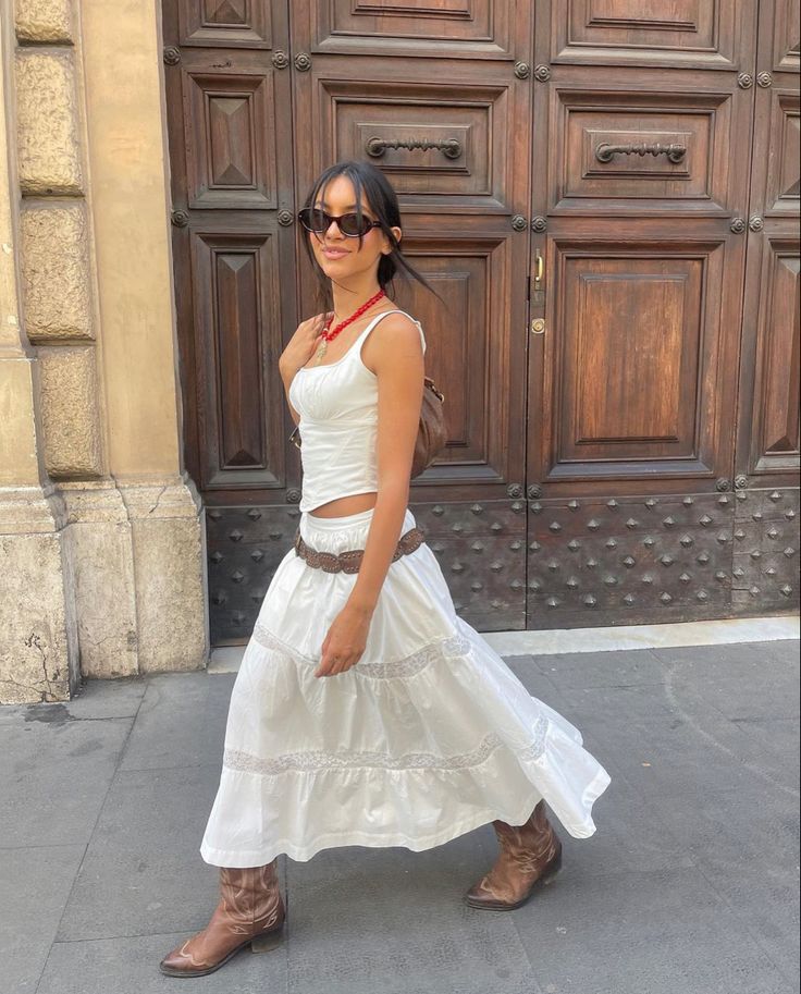 A woman wearing a white camisole and a flowing white maxi skirt, paired with brown cowboy boots, poses cheerfully in front of a large wooden door.