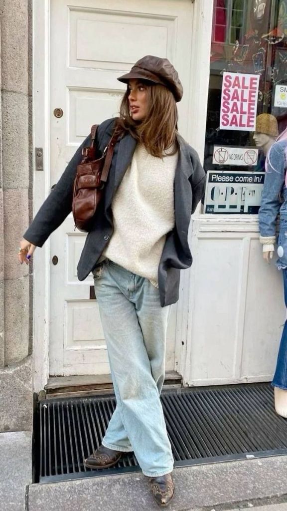A woman poses stylishly outside a store, wearing a comfortable oversized sweater, a long coat, wide-leg jeans, and a fashionable hat. She carries a brown bag and stands on a textured mat, with store signage visible in the background.