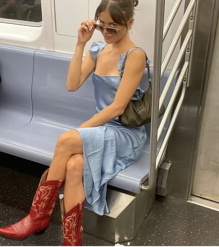 A person sitting on a subway bench, wearing a light blue dress and red cowboy boots, adjusting their sunglasses.