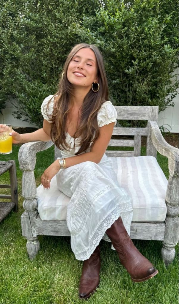A person sitting on a wooden bench in a garden, wearing a white dress and brown cowboy boots, smiling while holding a drink.