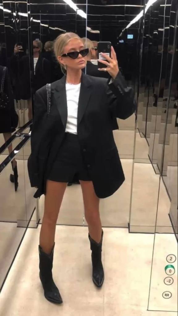 A woman taking a selfie in a mirror wearing a black oversized blazer, a white t-shirt, black shorts, and black cowboy boots, with sunglasses and a stylish handbag.