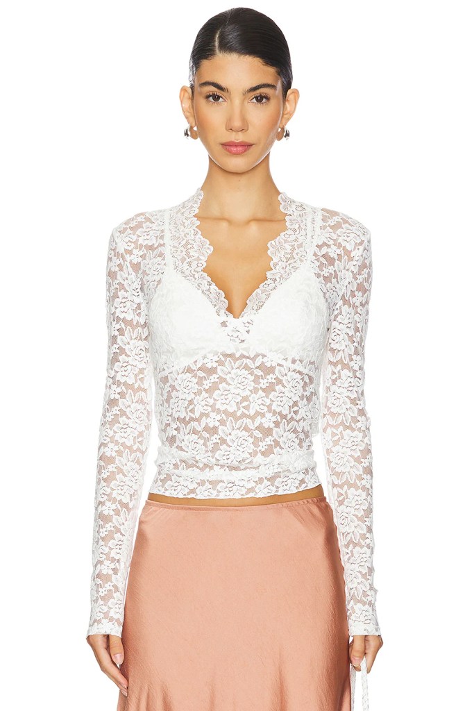 A woman modeling a white lace top with long sleeves, featuring a floral pattern and a deep neckline, paired with a soft pink satin skirt.