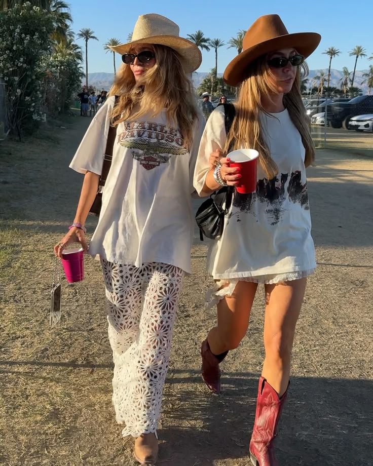 Two women walking outdoors, dressed in oversized graphic tees and stylish hats. One woman wears a pair of white lace pants and cowboy boots, while the other wears denim shorts with cowboy boots. They are holding drinks and enjoying a sunny day.
