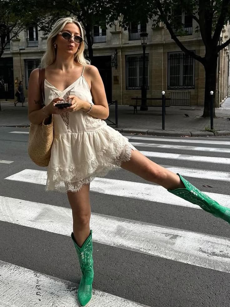 A woman wearing a light-colored mini dress and green cowboy boots poses playfully on a crosswalk.